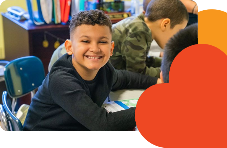 A young student smiles brightly while sitting at a classroom table, surrounded by other children working on activities. The classroom setting includes books, chairs, and colorful folders in the background.
