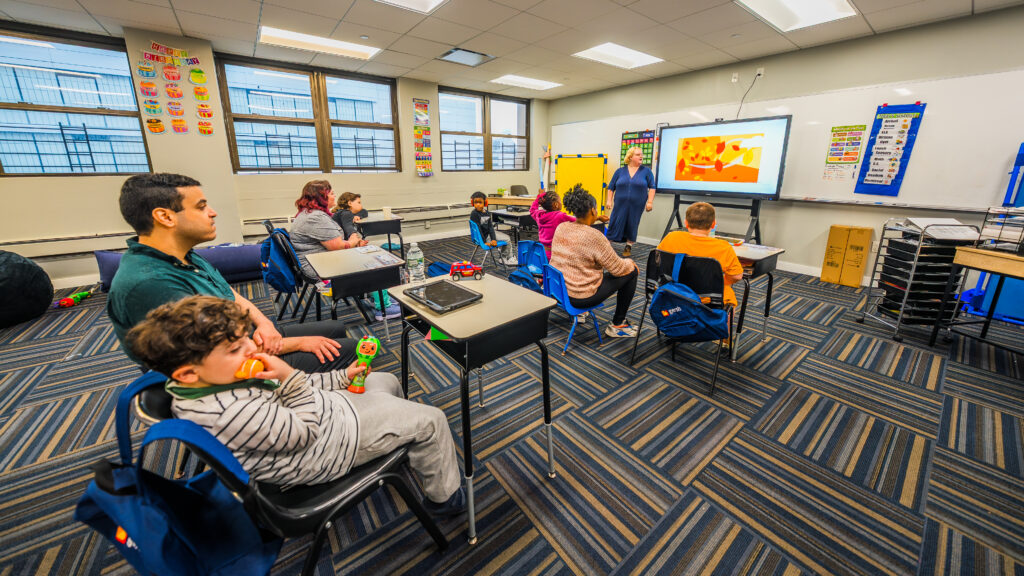 Students and teachers engaged in a classroom lesson at Gersh Autism Academy. Children sit at desks and on chairs while a teacher leads an interactive activity on a smartboard. The autism-specific learning environment fosters focus, communication, and social skills in a supportive classroom setting.