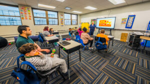 Students and teachers engaged in a classroom lesson at Gersh Autism Academy. Children sit at desks and on chairs while a teacher leads an interactive activity on a smartboard. The autism-specific learning environment fosters focus, communication, and social skills in a supportive classroom setting.
